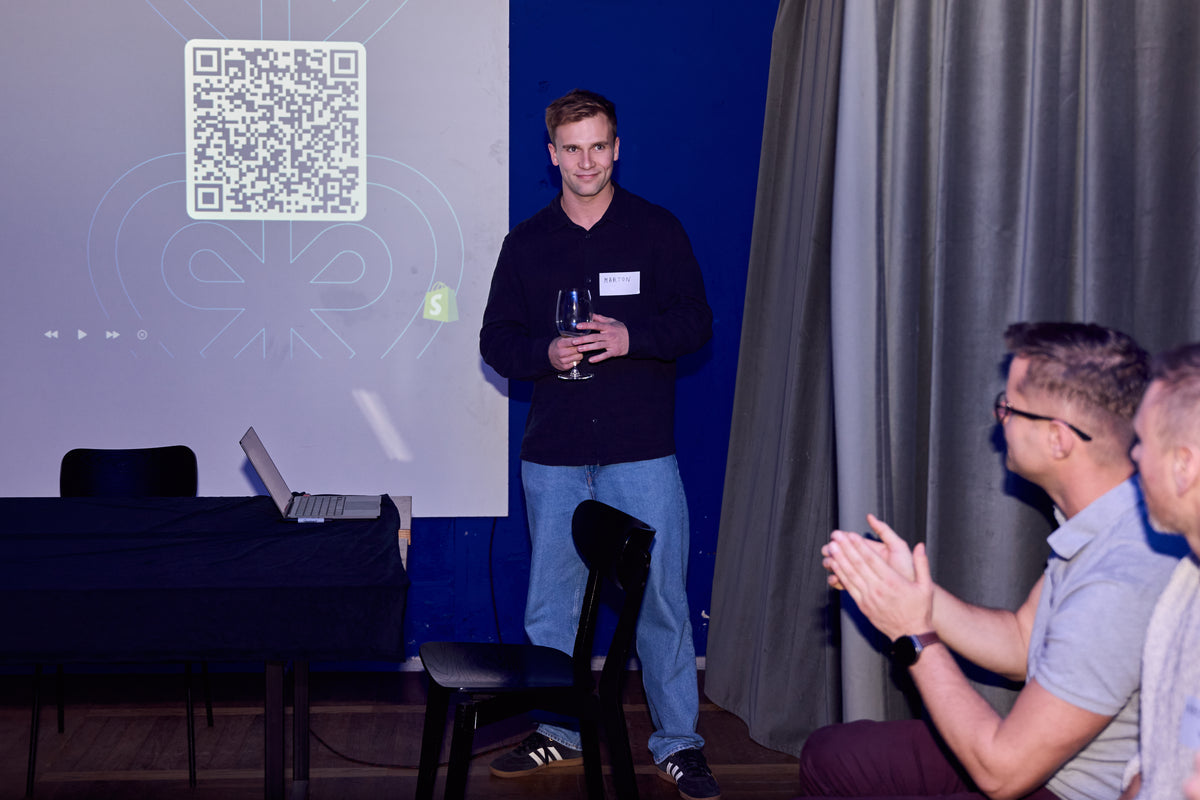 A man in a black long-sleeve shirt and blue jeans stands on a stage holding a wine glass, with a QR code projected on a screen behind him. Two seated audience members are clapping on the right, and a table with a laptop is visible on the left.