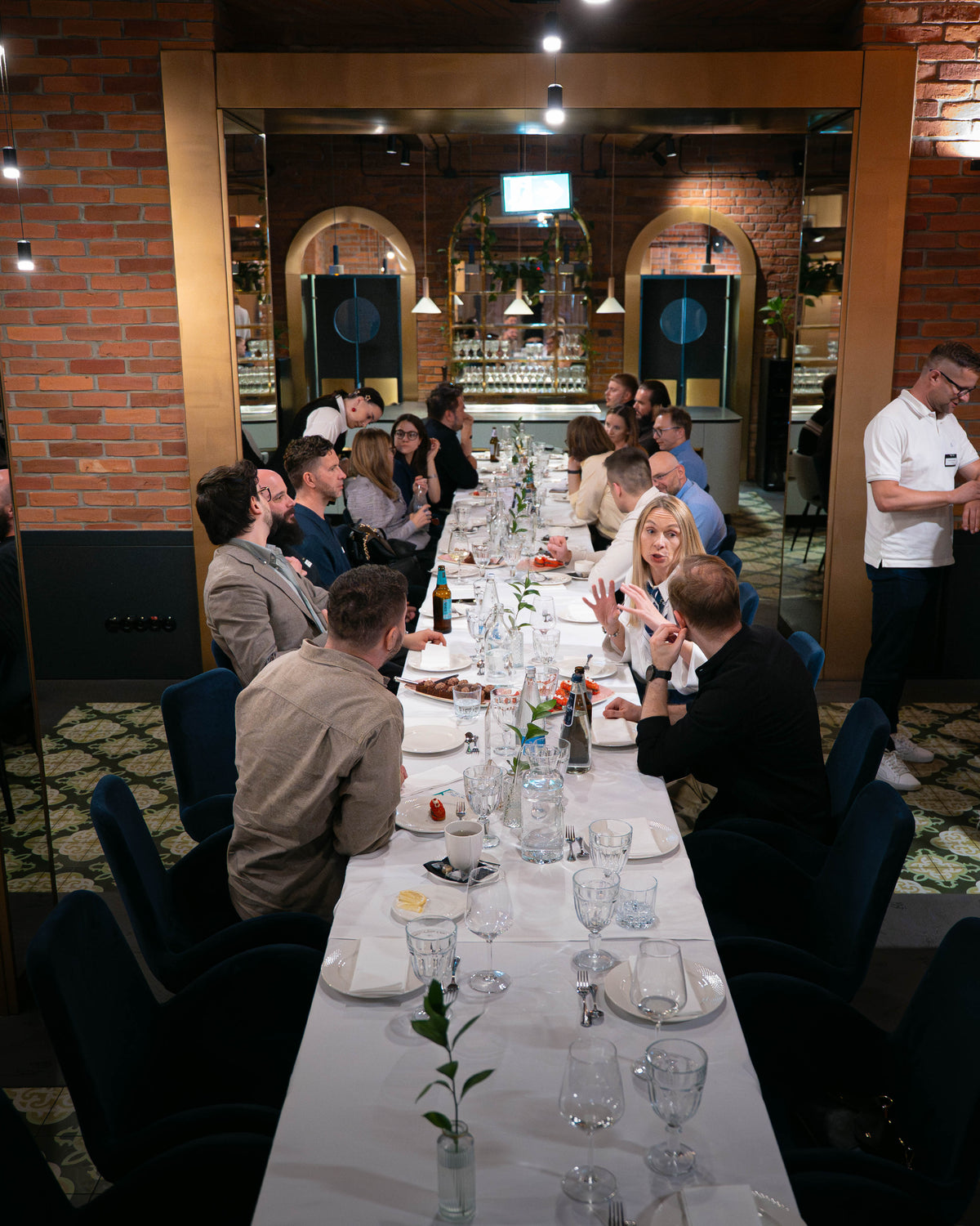A long dining table set in a restaurant with multiple people seated and standing, featuring brick walls, hanging lights, and a warm indoor ambiance.
