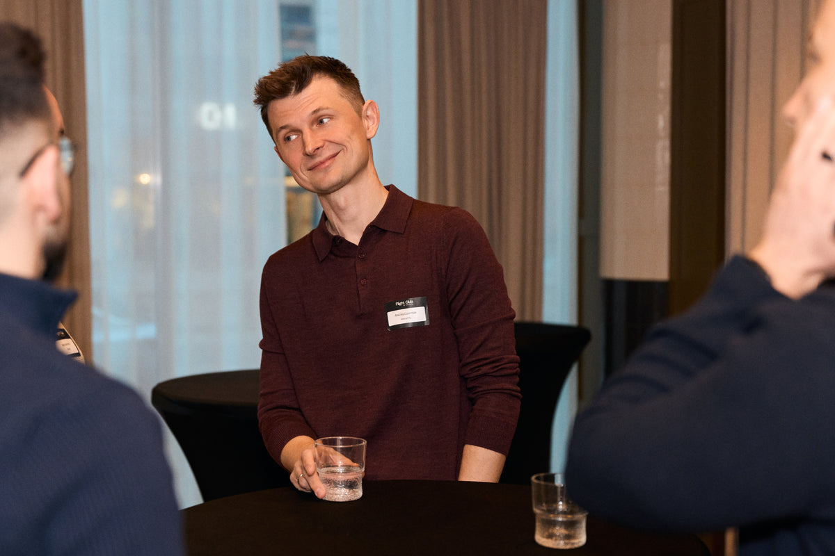 A man wearing a dark maroon long-sleeve polo shirt with a name tag sits at a table holding a glass of water, surrounded by two partially visible people in a modern indoor setting.