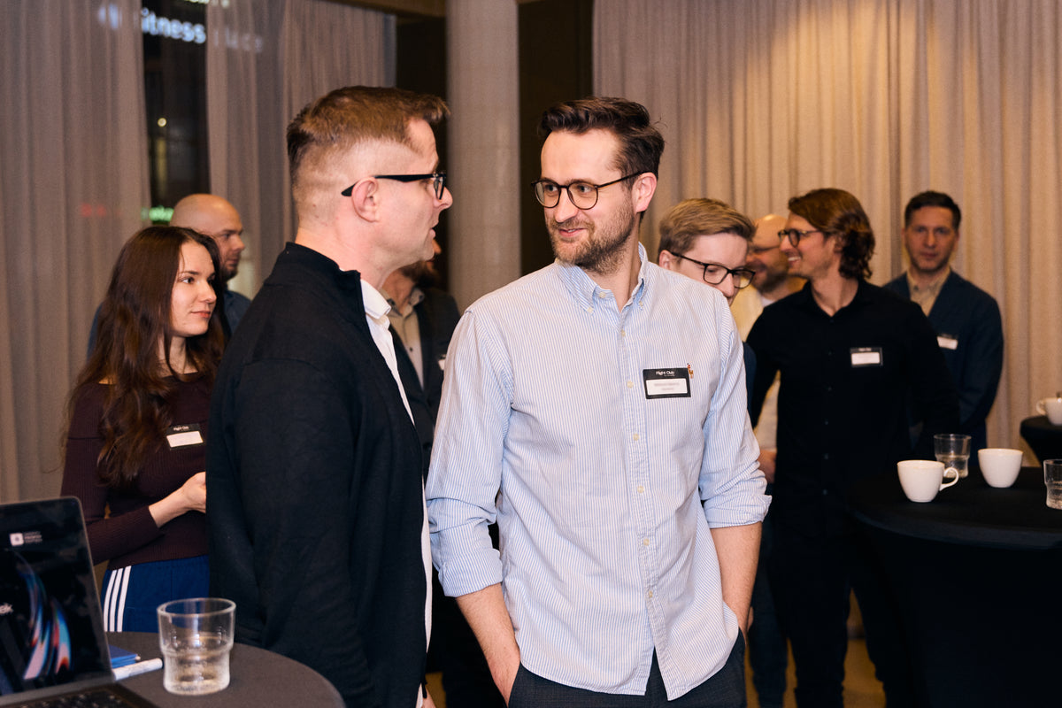 Two men in a networking event or conference setting, with one wearing a light blue button-down shirt and the other in a black blazer. Other attendees and tables with cups are visible in the background.