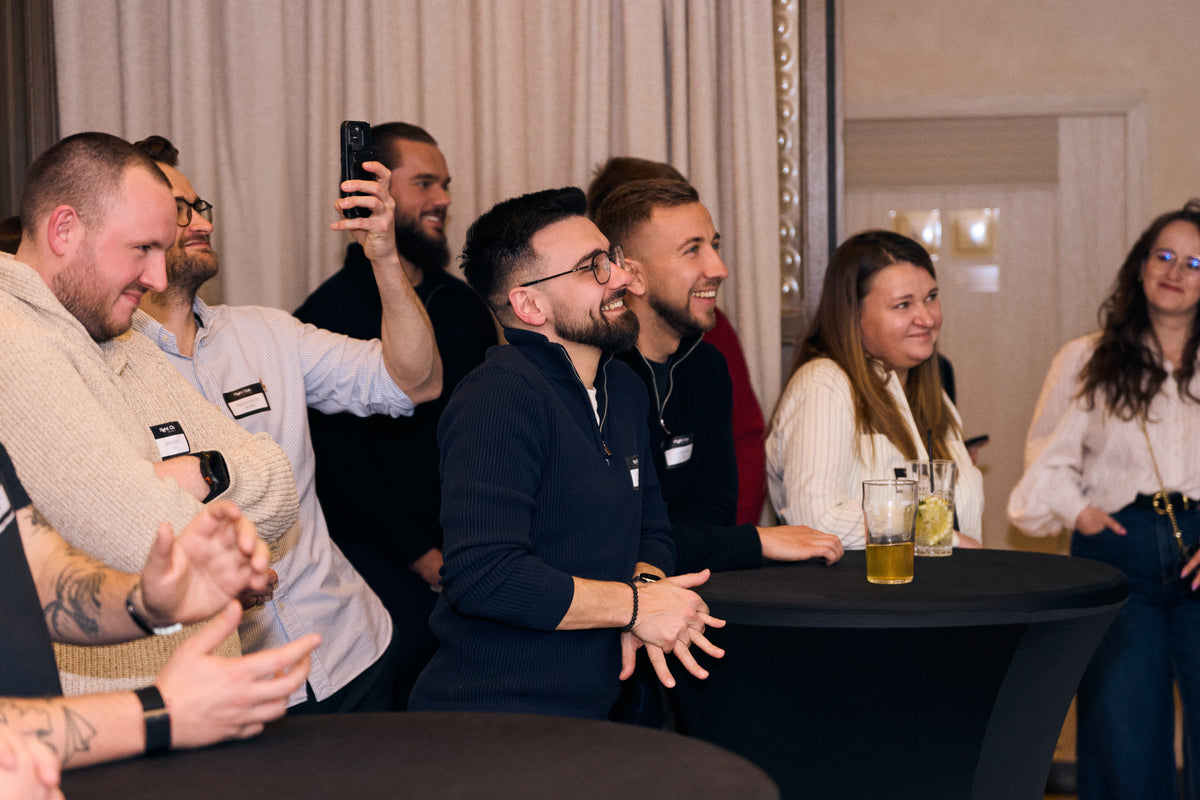 A group of people gathered around a black table in an indoor event space, with drinks on the table and a man in a dark navy hoodie at the center of the group.