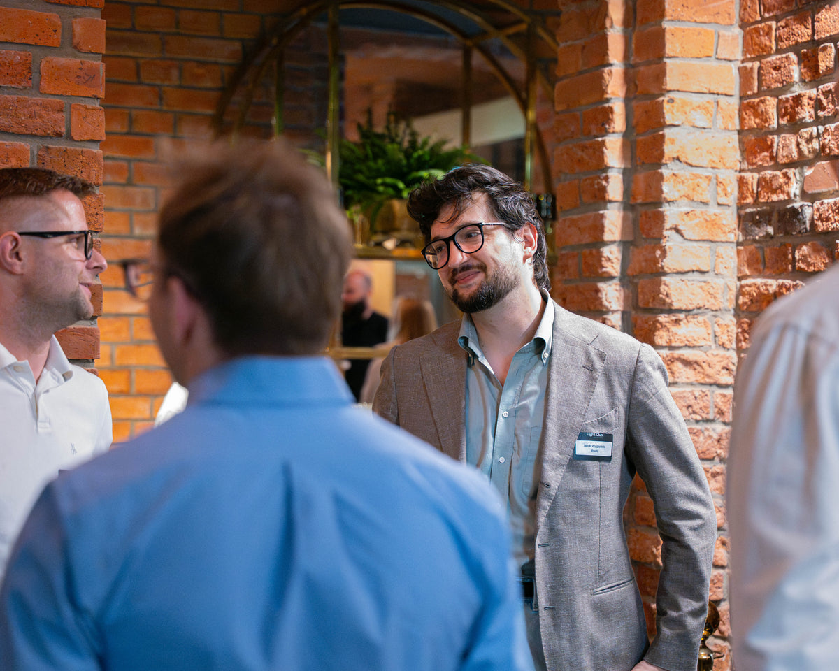 A man in a gray blazer and glasses wearing a name tag, standing in a brick-walled indoor setting and talking to people in the foreground.