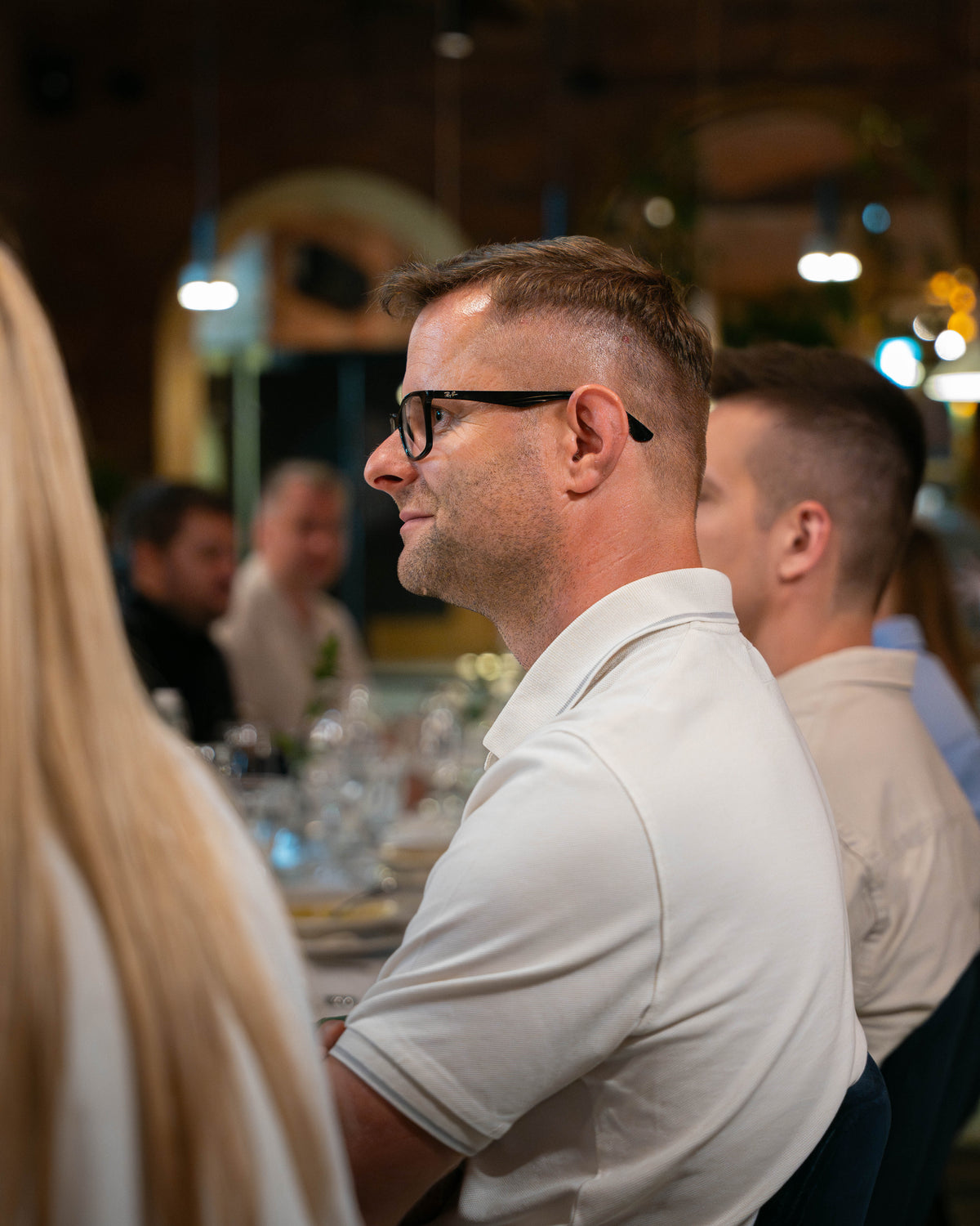 A man with short brown hair and black-rimmed glasses, wearing a white polo shirt, seated in a restaurant or event setting with other people blurred in the background.