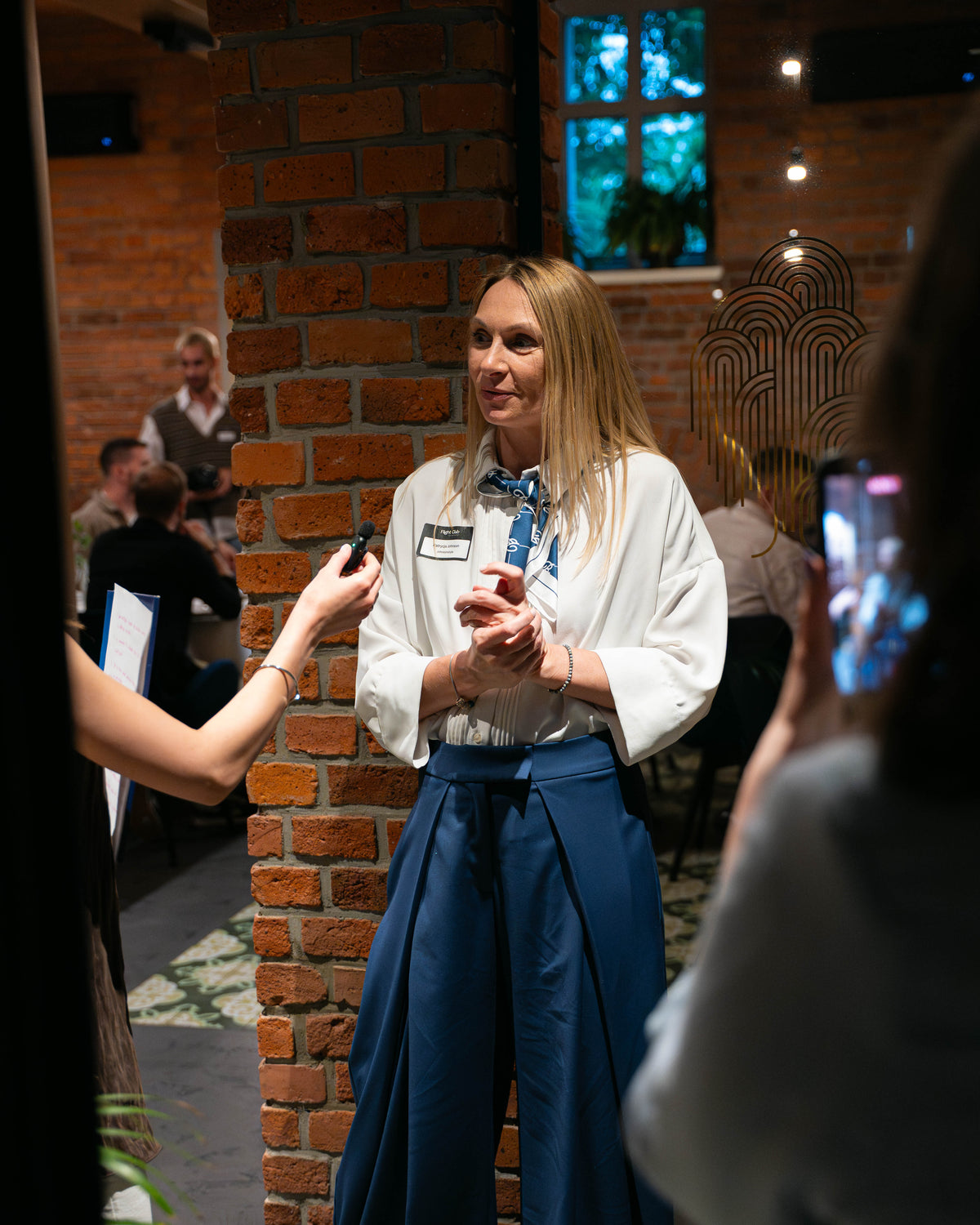 A woman in a white blouse and blue skirt stands indoors near a brick column, holding a small black object while being interviewed by someone holding a microphone. A phone is visible in the foreground, and other people and a restaurant-like interior are in the background.