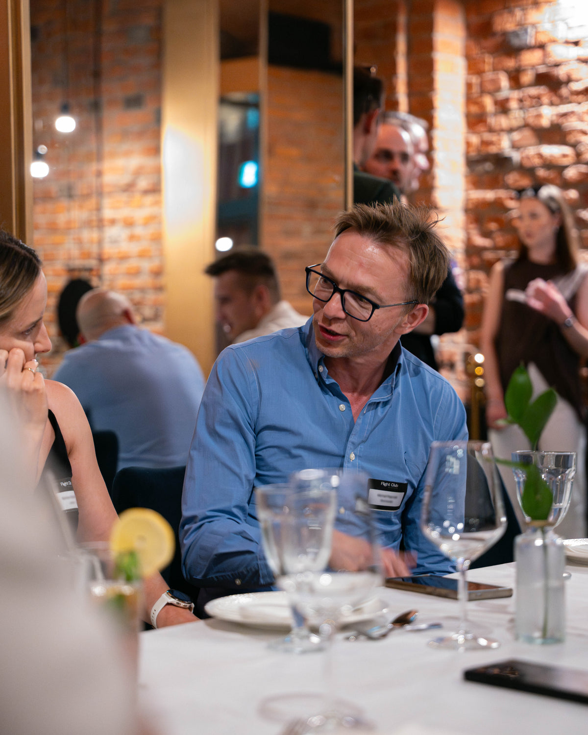 A man wearing a light blue button-down shirt and black-rimmed glasses sits at a restaurant table, engaged in conversation with a woman. The setting features a warm brick wall background, dining tables with wine glasses, and other diners in a softly lit indoor environment.