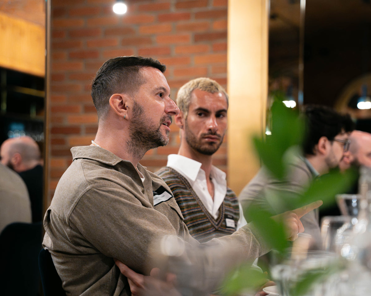 A man in a beige long-sleeve shirt sitting at a table in a restaurant or cafe, with another man in a striped sweater behind him and blurred foreground foliage.